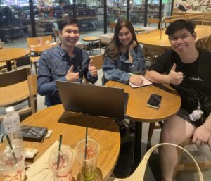 3 คนนั่งเรียนที่โต๊ะไม้ในร้านกาแฟพร้อมแล็ปท็อป