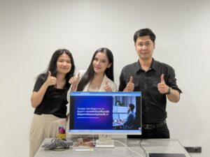 3 คนยืนข้างจอคอมพิวเตอร์ที่แสดงสไลด์ ชูนิ้วโป้ง