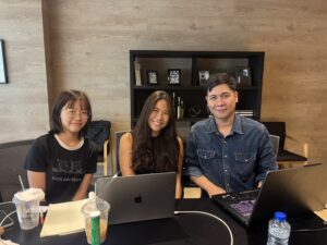 3 คนนั่งเรียนที่โต๊ะพร้อมแล็ปท็อป หน้าชั้นวางของ