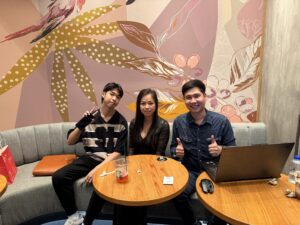 3 คนนั่งที่โต๊ะกลมพร้อมแล็ปท็อป หน้าผนังลายใบไม้
