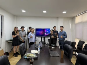 ผู้เรียนยืนรอบโต๊ะประชุมในห้องอบรมที่มีจอแสดงสไลด์